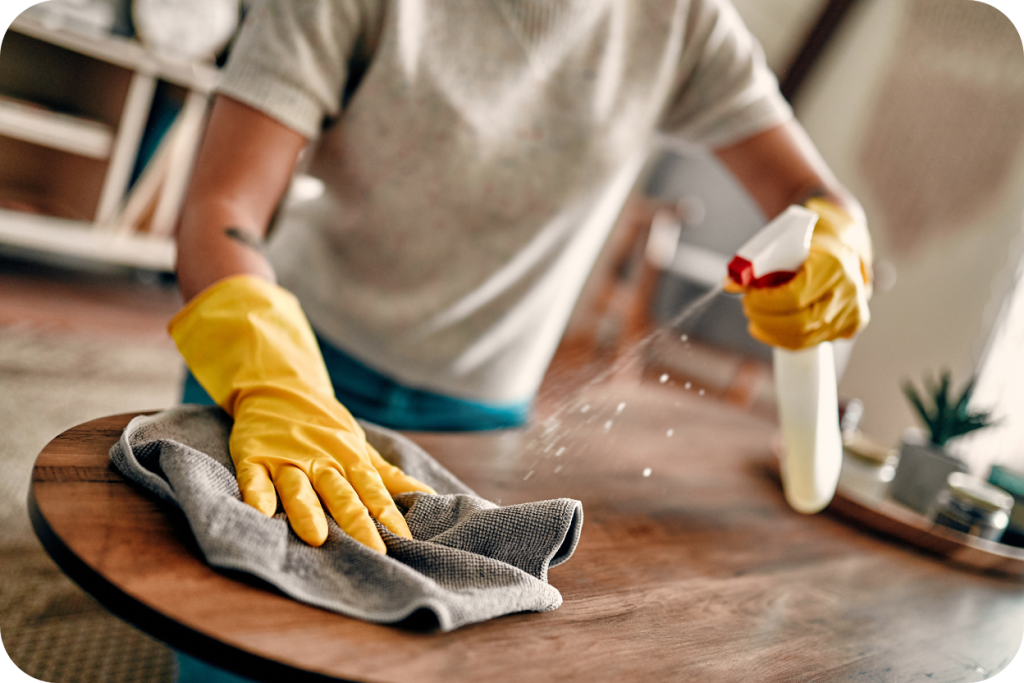 Professional home cleaner wearing bright yellow gloves sprays cleaner and wipes a wooden table with a microfiber cloth.